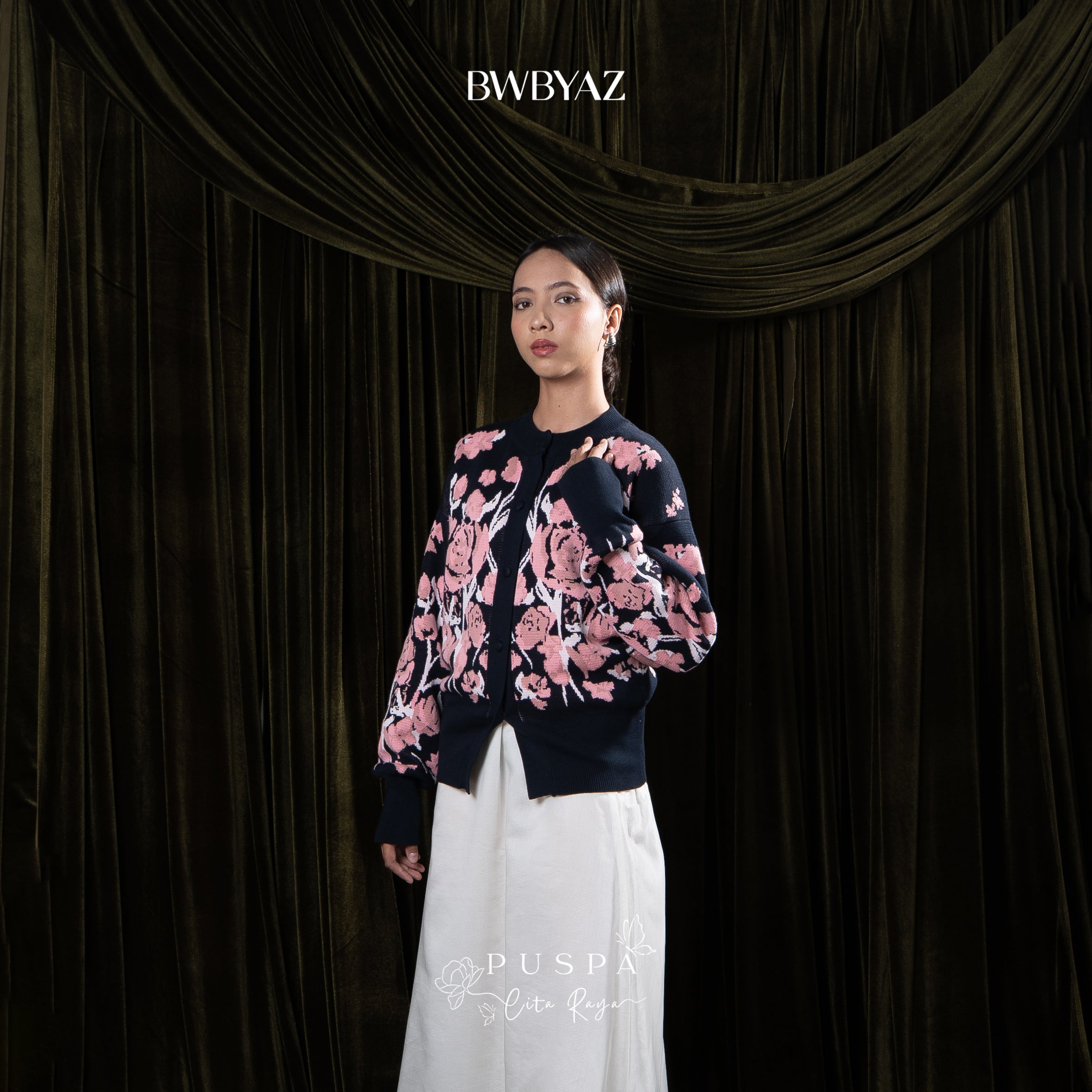 Woman wearing a navy floral cardigan and white skirt against a dark curtain background with 'BWBYAZ' branding.