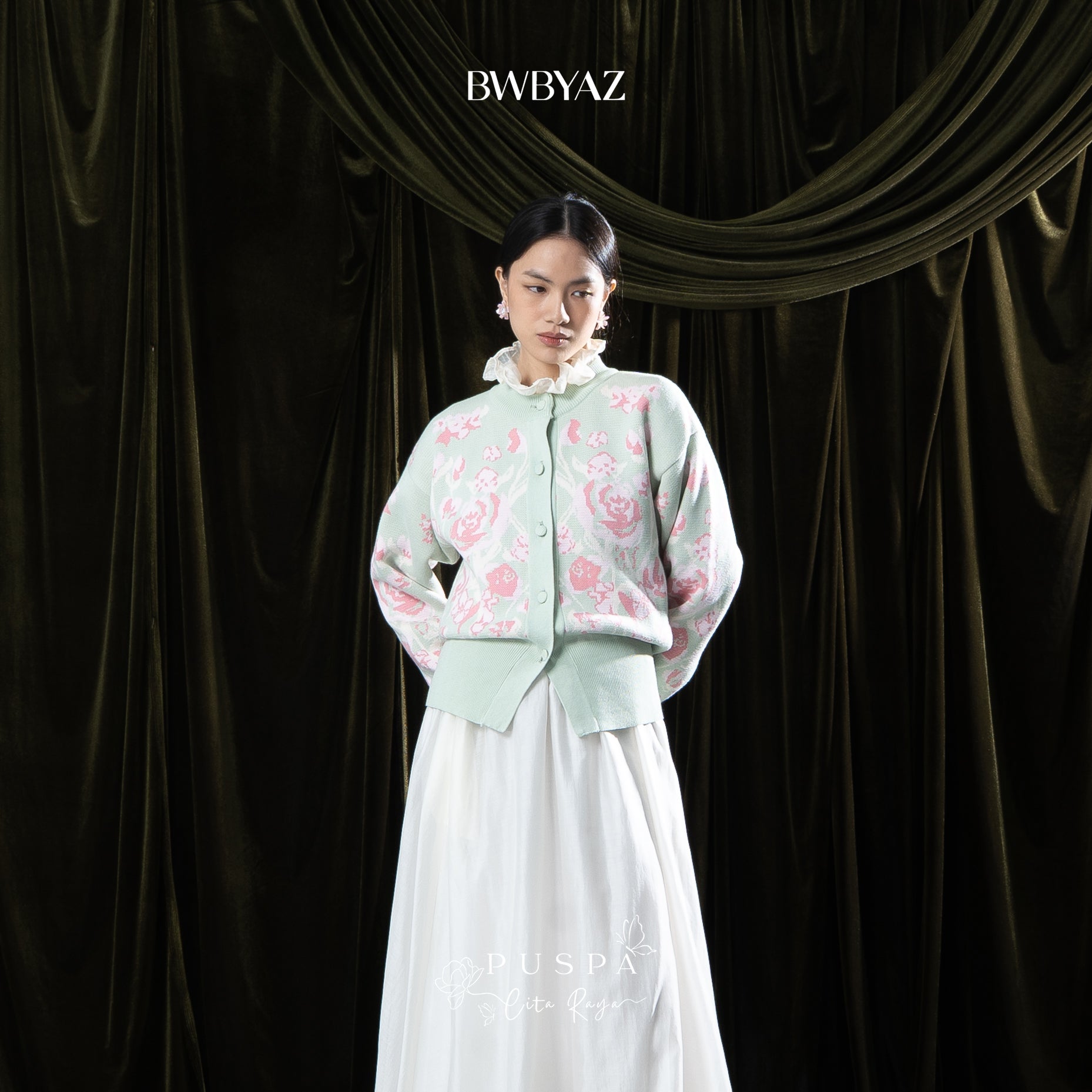 Woman wearing a light green floral cardigan with a white skirt against a dark curtain background, featuring the brand 'BWBYAZ'.