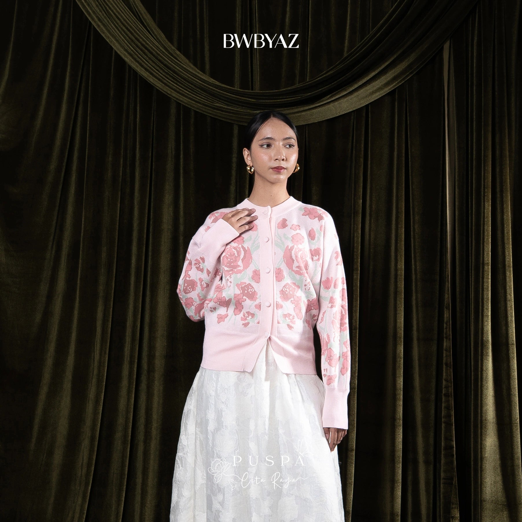 Woman wearing a pink floral cardigan and white skirt against a dark curtain background with 'BWBYAZ' branding.