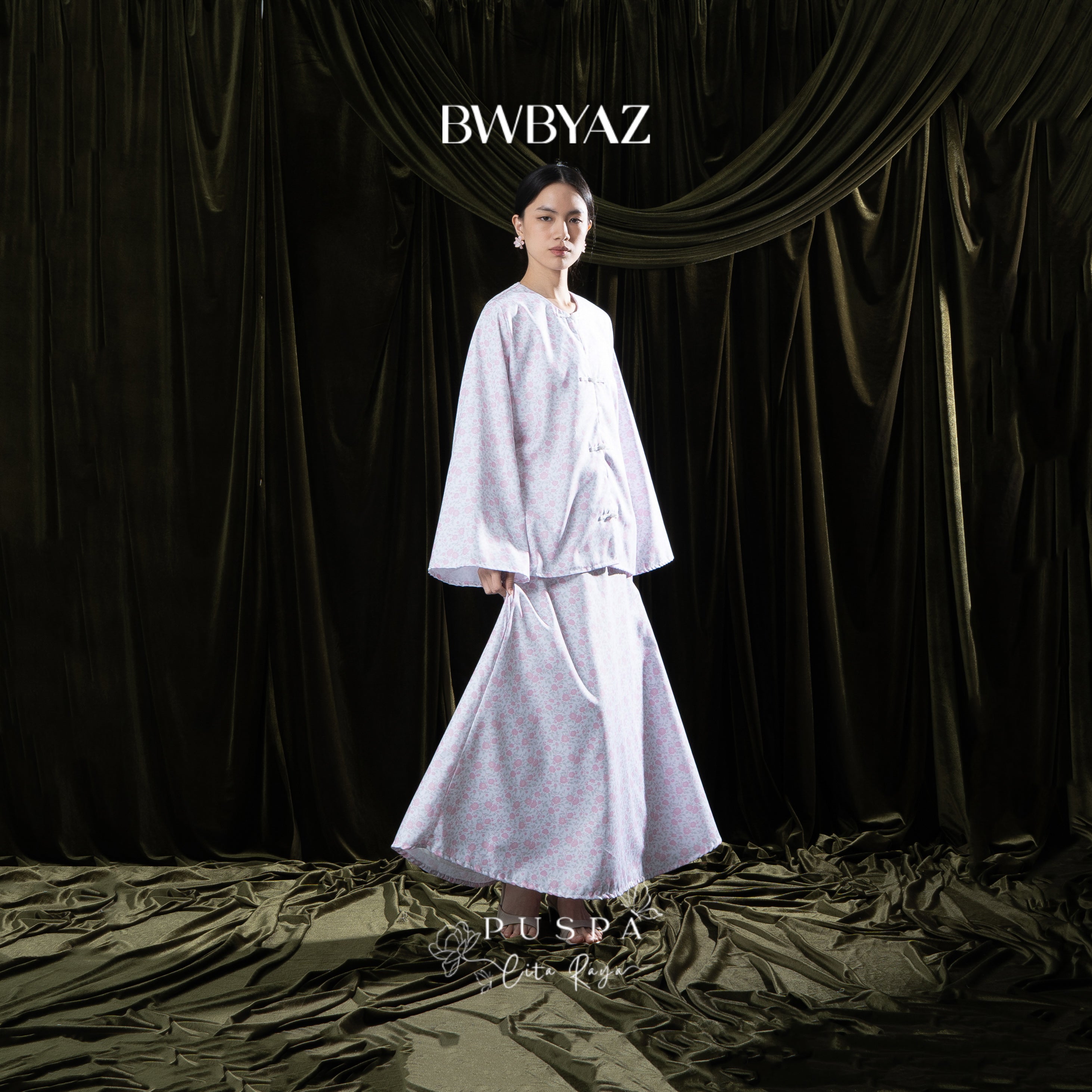 Person wearing a white floral skirt with 'BWBYAZ' branding against a dark curtain background.