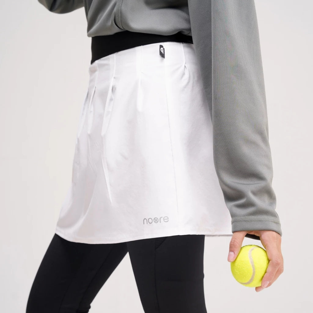 A person wearing a white tennis skirt by Noore with a logo on the lower left side, paired with black leggings and a gray sweatshirt. A tennis ball is held in the person's hand.