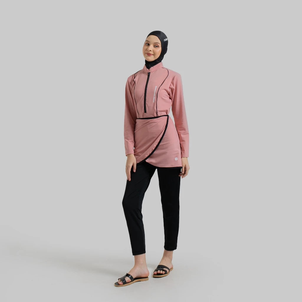 A woman wearing a dusty pink Serenity Burkini by Noore, consisting of a long-sleeved top, bottom, and a detachable skirt, paired with black pants.