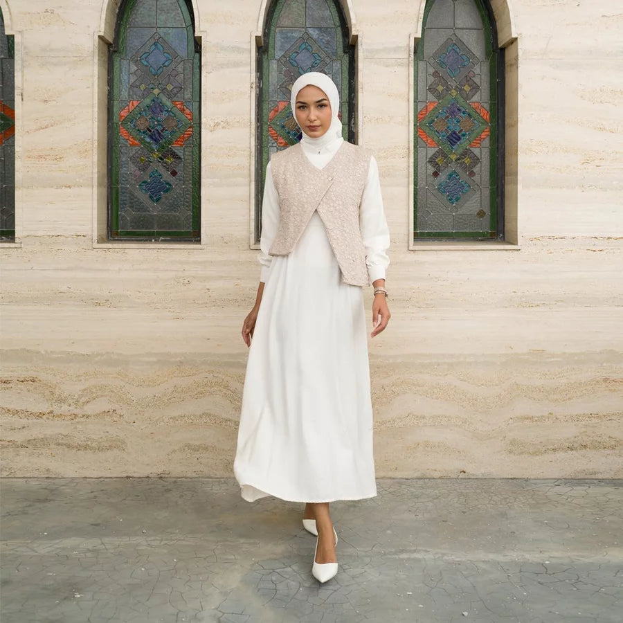 Woman in a white dress by Tunique with a beige vest standing in front of a building with stained glass windows.