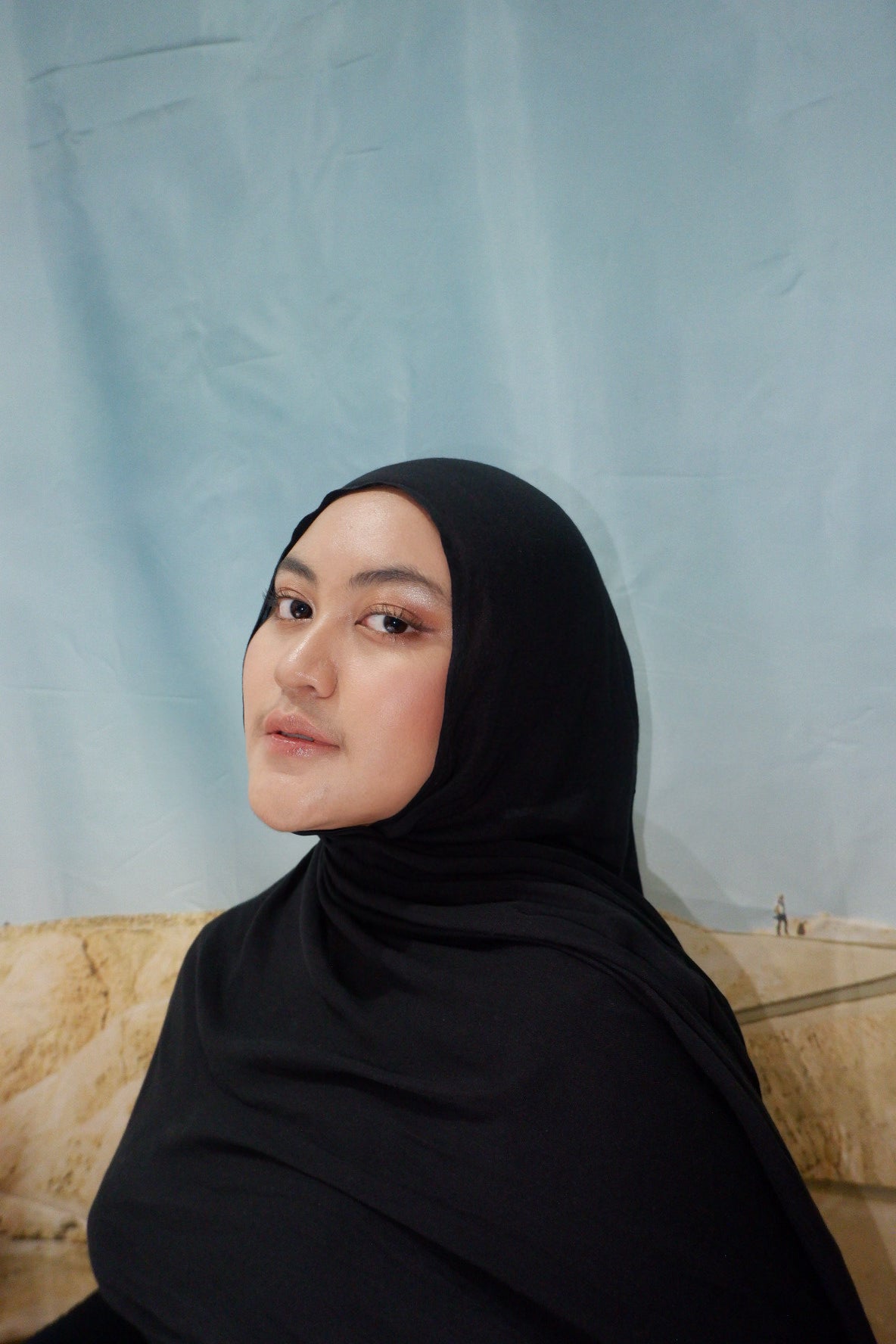 Woman wearing a black pashmina viscose sitting on a beige couch with a light blue curtain background