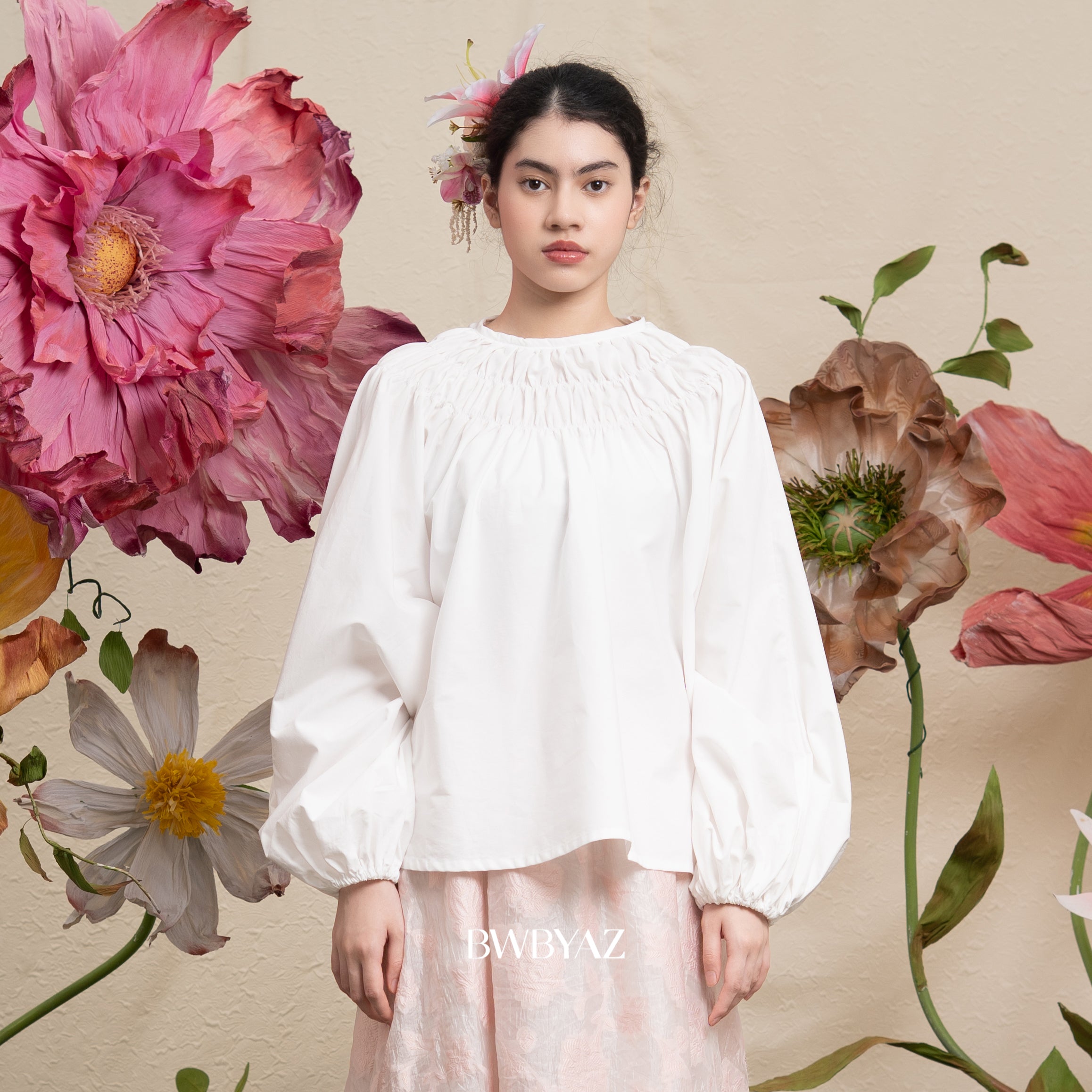 Woman in a ivory white blouse standing against a floral background with 'BWBYAZ' branding.