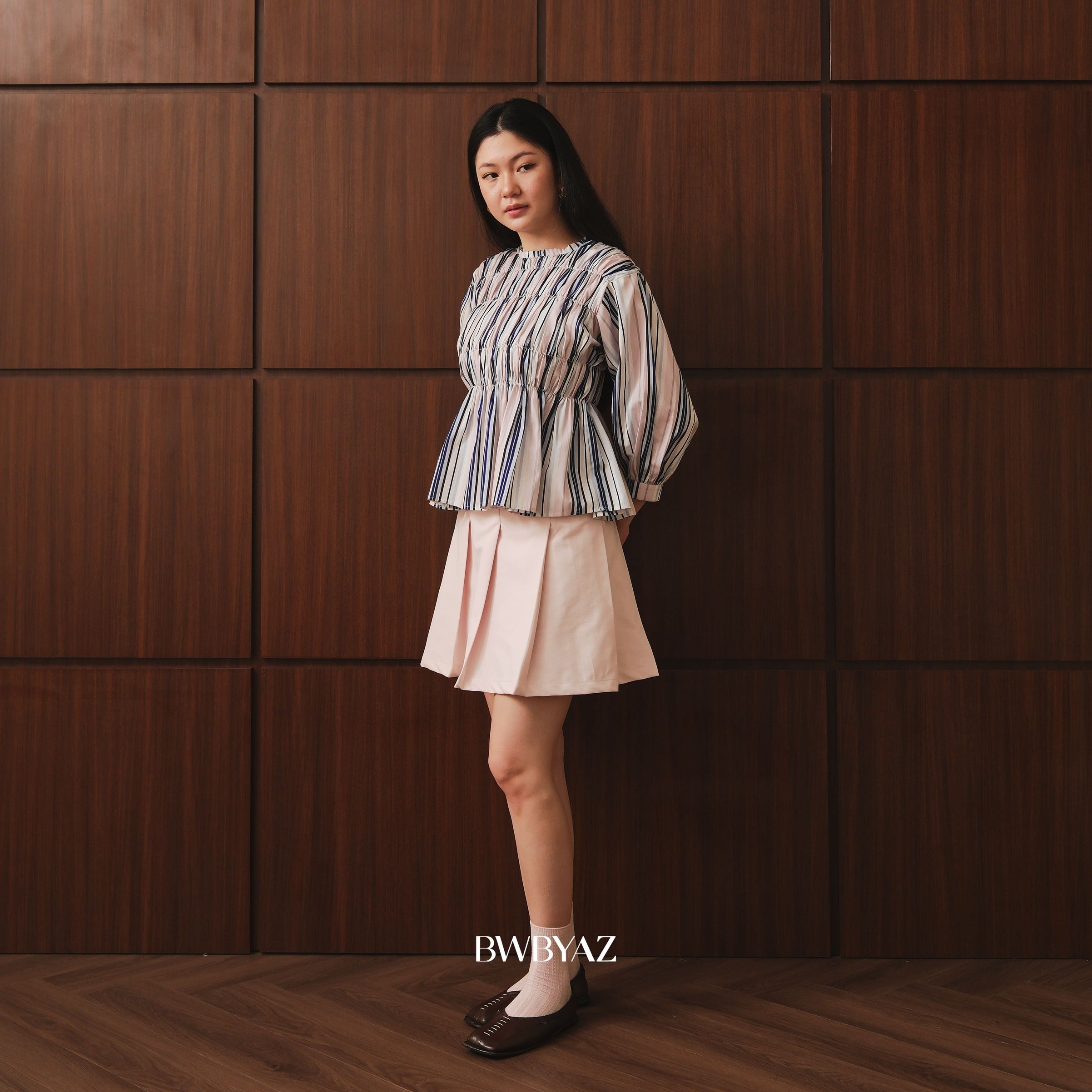 A woman stands against a dark brown wood-paneled wall wearing a striped long-sleeve peplum top by BWBYAZ and a light pink pleated mini skirt. She is wearing white socks and dark brown flat shoes.