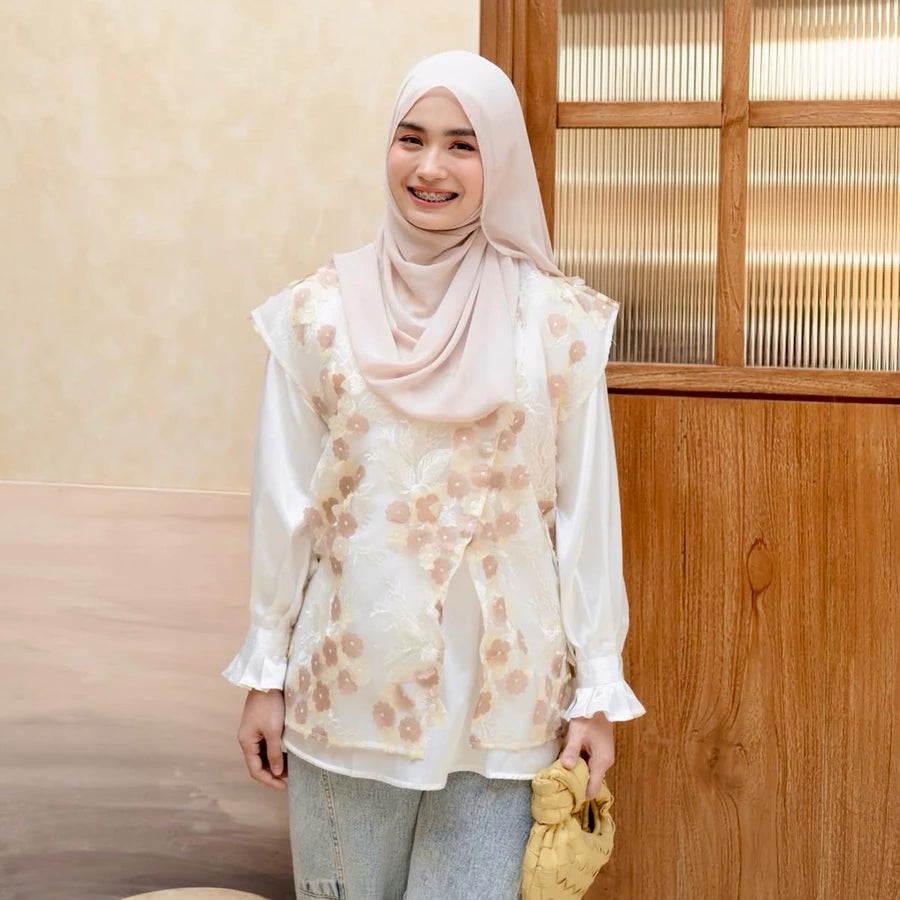 Woman wearing a floral vest by Haidee Orlin over white shirt and hijab standing indoors with a wooden panel background
