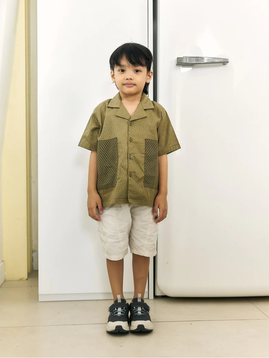 A young boy stands in front of a white refrigerator wearing a short-sleeve olive green button-up shirt by Studio Tui with textured front panels, off-white shorts, and dark sneakers.