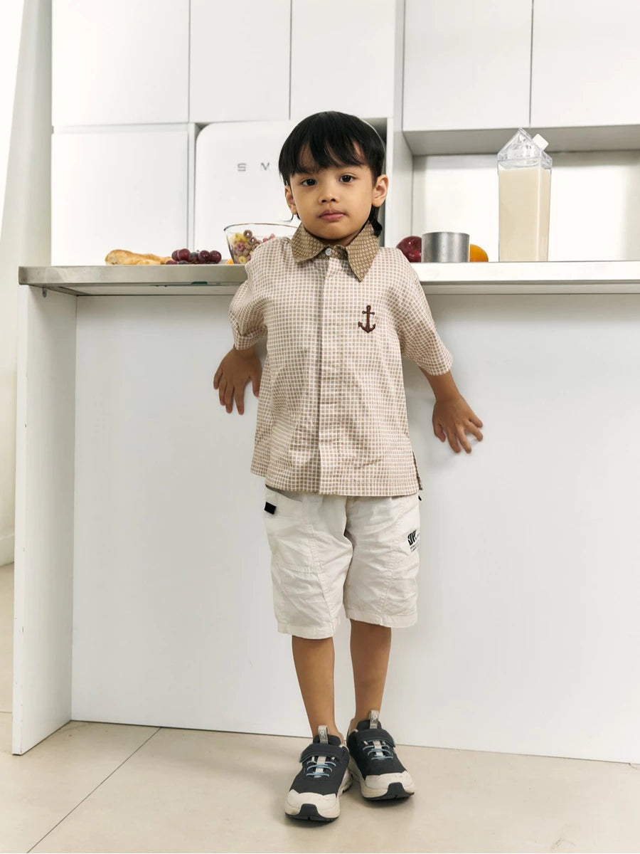 A young boy stands in a bright, modern kitchen wearing a beige and white checkered short-sleeve button-up shirt by Studio Tui with a brown collar and a small anchor emblem, paired with off-white shorts and dark sneakers.
