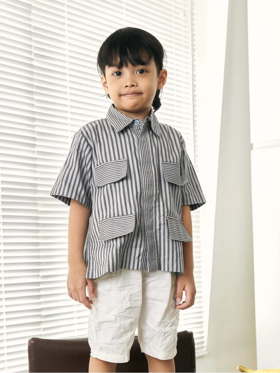 A young boy wearing a short-sleeve button-up shirt by Studio Tui with vertical gray and white stripes, featuring large front flap pockets, paired with off-white shorts, standing in front of white window blinds.