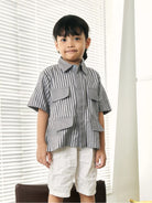 A young boy wearing a short-sleeve button-up shirt by Studio Tui with vertical gray and white stripes, featuring large front flap pockets, paired with off-white shorts, standing in front of white window blinds.