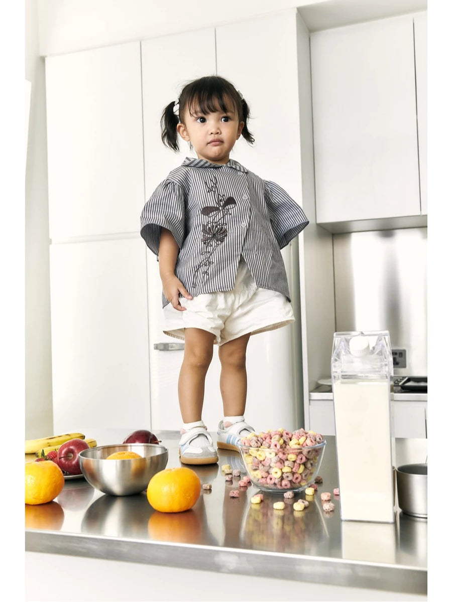 A toddler girl stands in a modern white kitchen wearing a gray and white striped smock top by Studio Tui with a dark floral print, white shorts, and white sneakers. In the foreground on a stainless steel counter are bowls of fruit, cereal, and a carton.