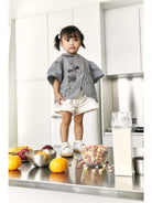 A toddler girl stands in a modern white kitchen wearing a gray and white striped smock top by Studio Tui with a dark floral print, white shorts, and white sneakers. In the foreground on a stainless steel counter are bowls of fruit, cereal, and a carton.