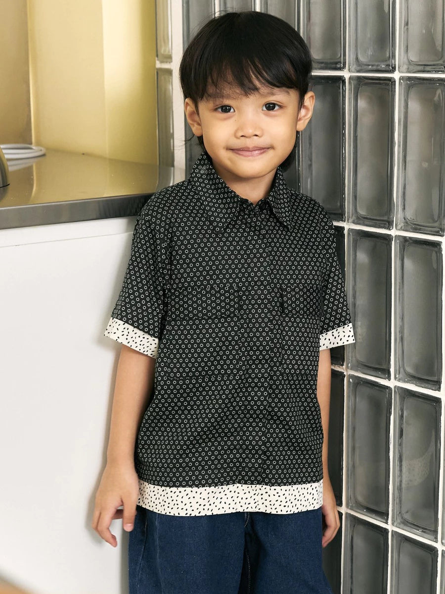 A young boy wearing a short-sleeve black button-up shirt by Studio Tui with a white geometric pattern and contrasting white cuffs and hem trim, paired with dark blue jeans, standing in front of a glass block wall.