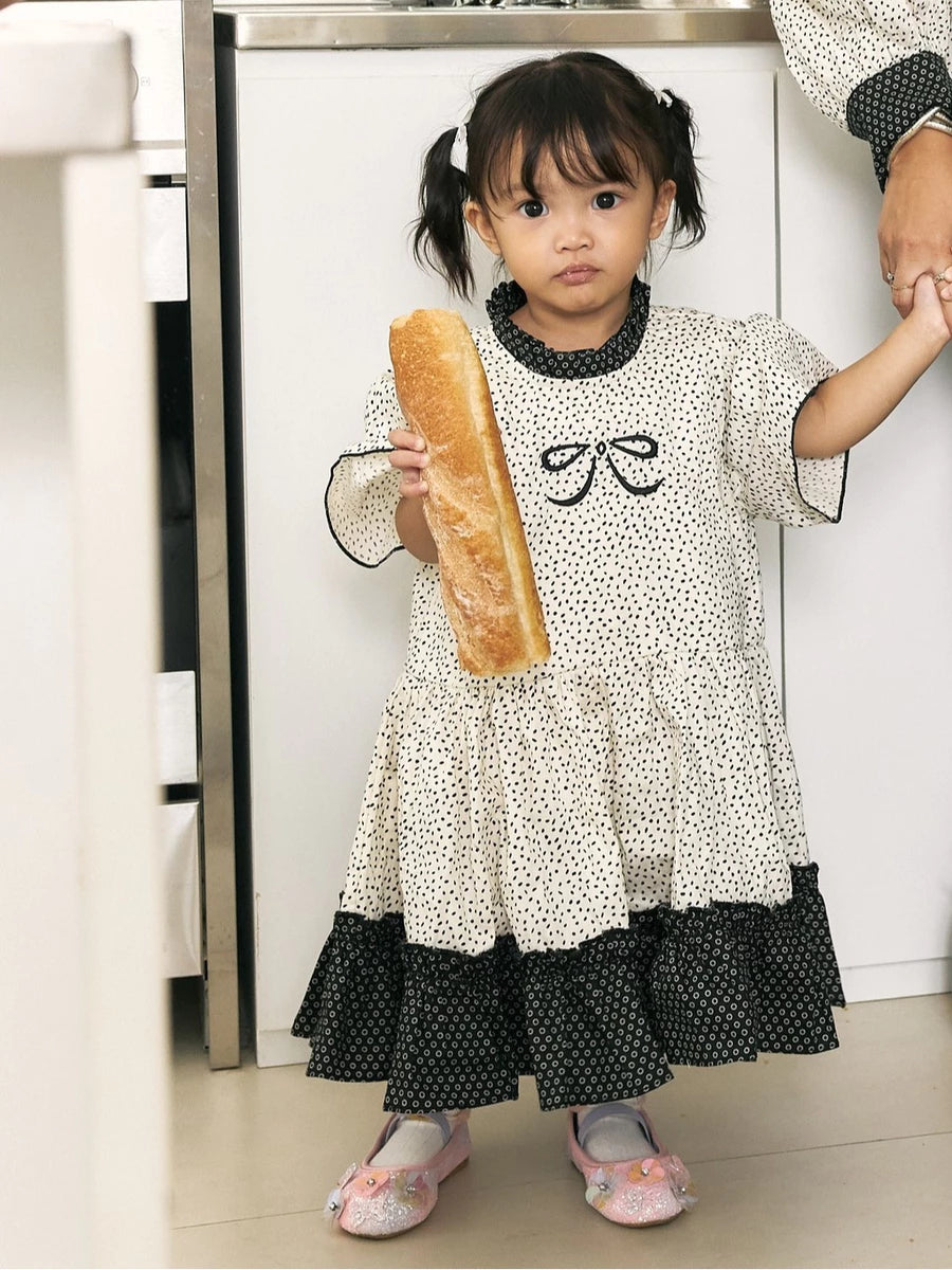 A young girl with dark hair in pigtails stands indoors holding a baguette. She is wearing a cream-colored dress by Studio Tuiwith black polka dots, black ruffled trim at the collar and hem, and a black bow motif on the chest.