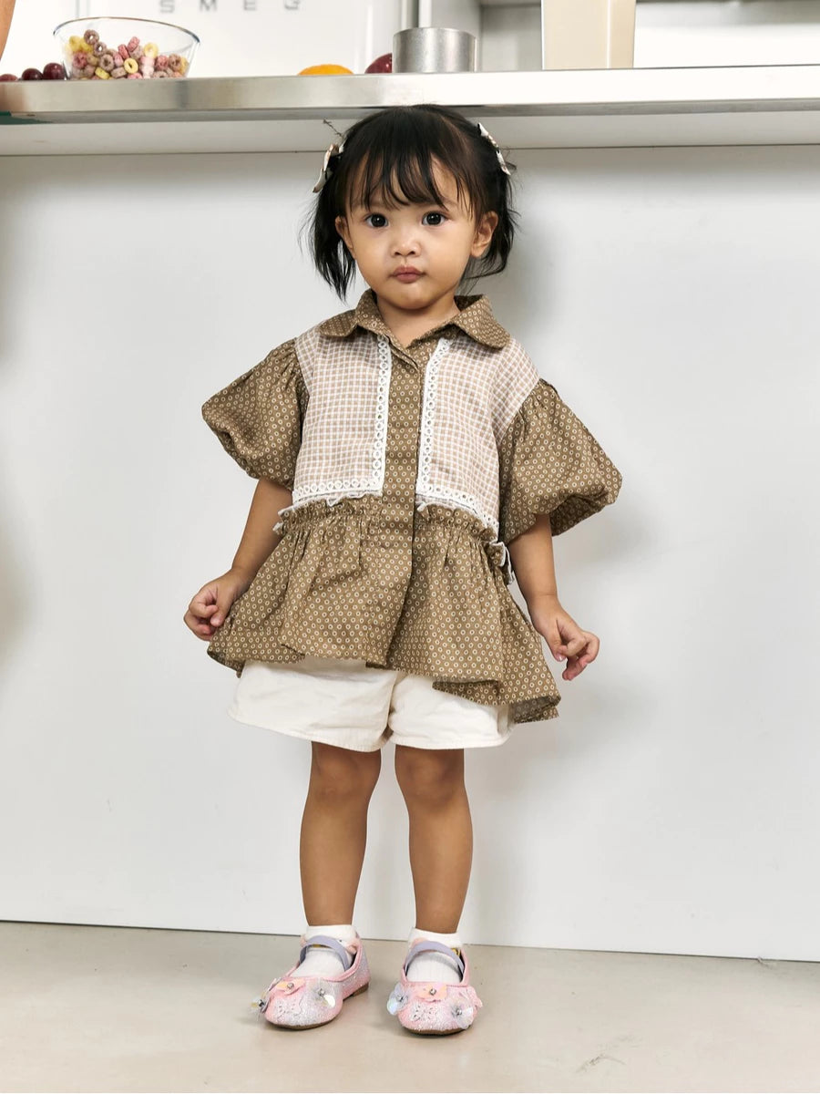 A toddler girl wearing a brown and beige patterned short-sleeve top by Studio Tui with white shorts and pink shoes, standing in front of a white wall.