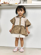 A toddler girl wearing a brown and beige patterned short-sleeve top by Studio Tui with white shorts and pink shoes, standing in front of a white wall.