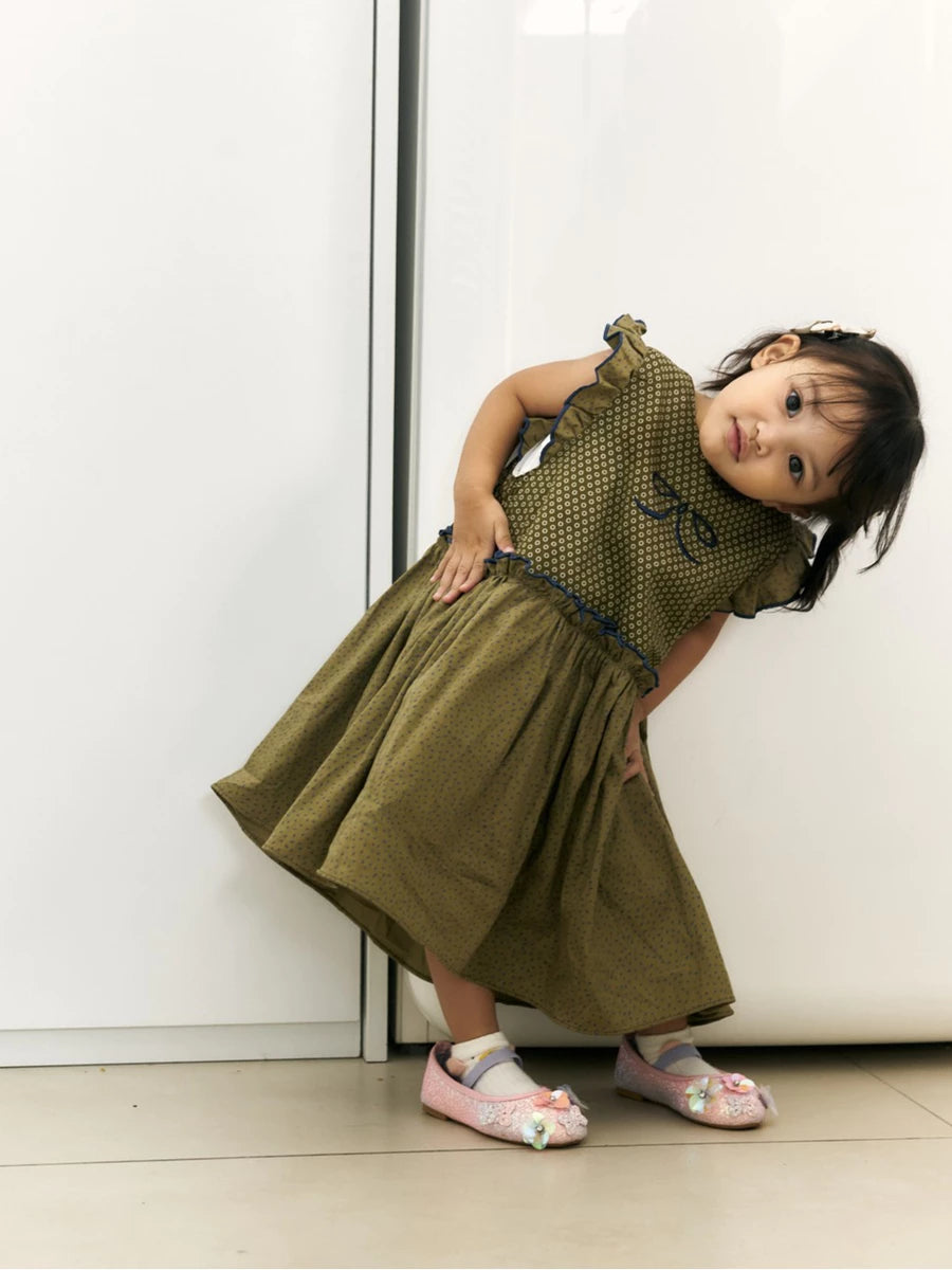 A toddler girl wearing an olive green sleeveless dress by Studio Tui with a polka dot bodice, ruffled shoulders, and a flared skirt, posing against a white wall.