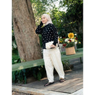 A woman wearing a black long-sleeve sweater by Haidee Orlin with white polka dots and cream-colored wide-leg trousers, standing on a wooden deck next to a large tree trunk with a green bench and a basket of flowers in the background.