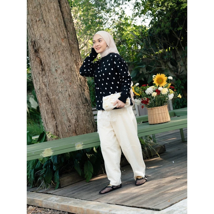 A woman wearing a black long-sleeve sweater by Haidee Orlin with white polka dots and cream-colored wide-leg trousers, standing on a wooden deck next to a large tree trunk with a green bench and a basket of flowers in the background.