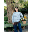 A woman stands outdoors in a garden setting wearing a light blue crewneck sweatshirt by Haidee Orlin with a small brown polka dot pattern, paired with dark blue wide-leg jeans and a brown lace hem detail. She is holding a small floral handbag next to a green bench with a basket of flowers.