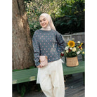 A woman wearing a dusty blue-gray long-sleeve knit sweater by Haidee Orlin with pink polka dots, a light beige hijab, and white lace-trimmed pants, standing outdoors next to a green bench with a flower basket.