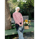 A woman wearing a pink long-sleeve sweater by Haidee Orlin with black polka dots, a white hijab, and light blue jeans stands outdoors on a green bench holding a black crescent handbag. A basket of flowers sits on the bench beside her, with trees and garden foliage in the background.