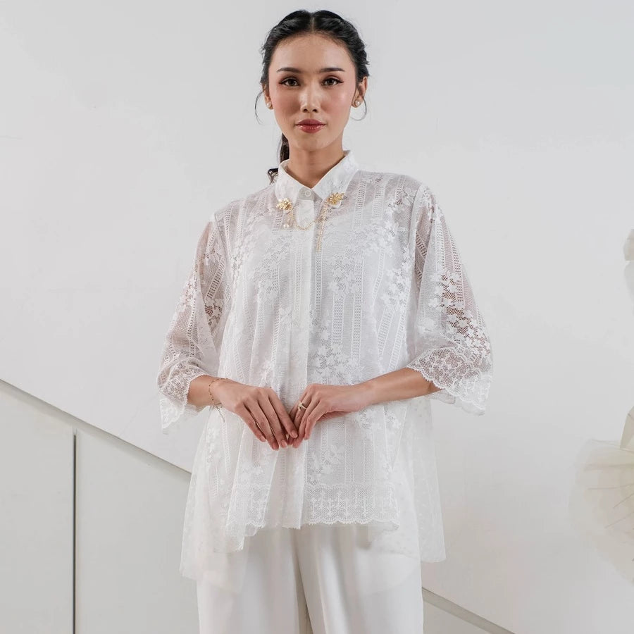 Woman wearing a white lace outer by Holy Peony and white pants against a light background
