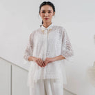 Woman wearing a white lace outer by Holy Peony and white pants against a light background