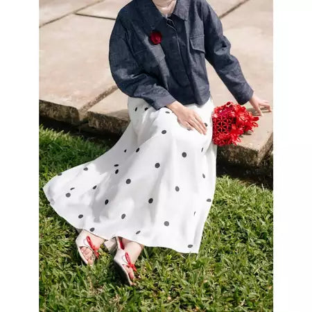 Person wearing a denim jacket and white polka dot skirt by Haidee Orlin holding red flowers on grass.