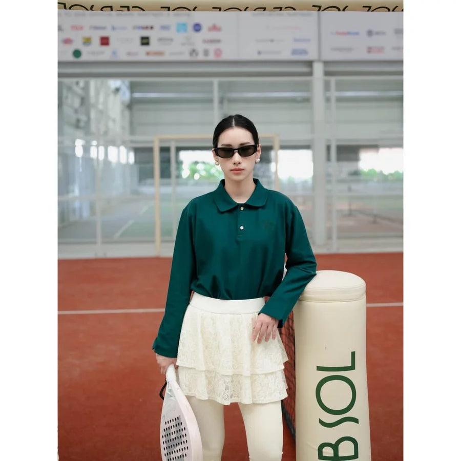 A woman stands on an indoor tennis court wearing an emerald green long-sleeve polo shirt by Haidee Orlin, a white tiered skirt, white tights, and black sunglasses, holding a tennis racket next to a BNSOL bollard.