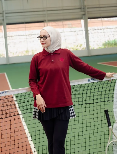 A woman wearing a hijab and a long-sleeve polo shirt in deep red by Haidee Orlin, standing on an indoor tennis court behind a net.