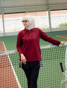 A woman wearing a hijab and a long-sleeve polo shirt in deep red by Haidee Orlin, standing on an indoor tennis court behind a net.