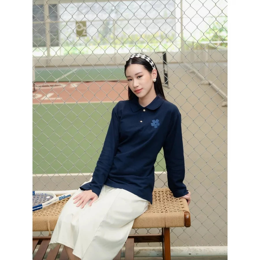 A woman sits on a woven bench in front of a chain-link fence at an outdoor sports field. She is wearing a navy blue long-sleeve polo shirt by Haidee Orlin with a small blue floral embroidery on the chest, paired with a cream-colored skirt.