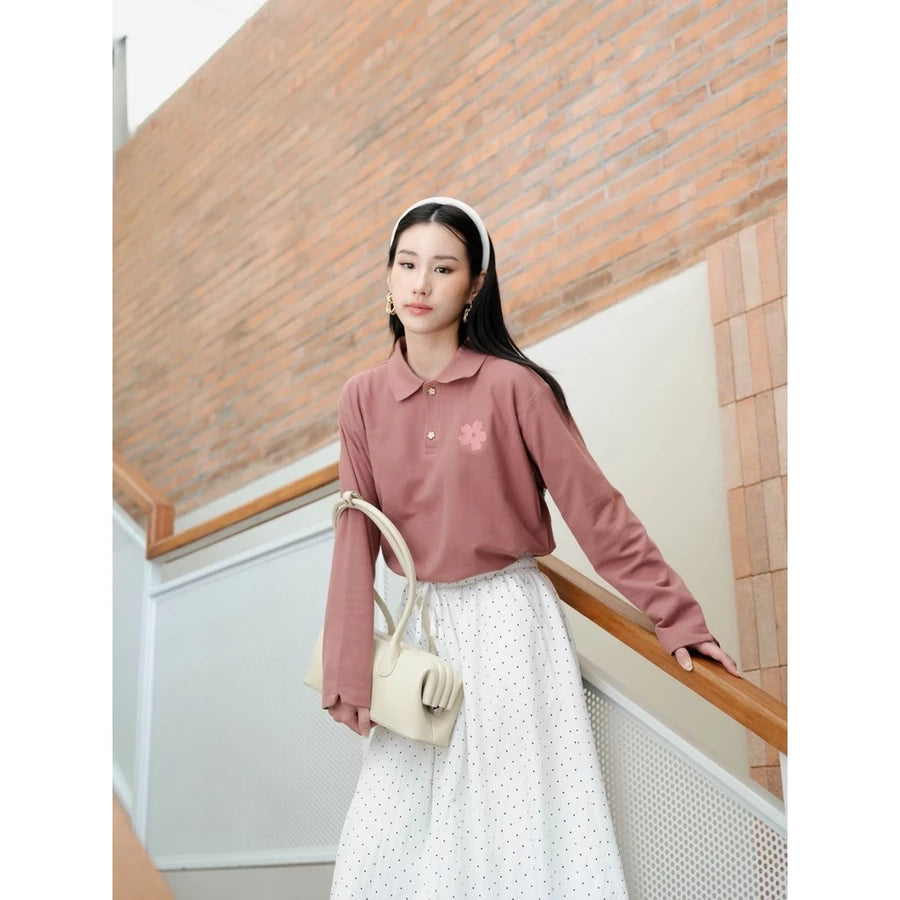 A woman stands on an indoor staircase wearing a dusty rose long-sleeve polo shirt by Haidee Orlin with a small pink flower detail on the chest, paired with a white polka-dot skirt and carrying a cream-colored shoulder bag.