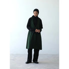 A person wearing a long green vest by Rashawl with a round neckline, sleeves, and a split at the back, paired with dark pants and black shoes.