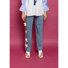 A person standing against a purple background, wearing blue denim pants with decorative white flowers on the leg, paired with a white shirt and beige shoes.

Seorang wanita berdiri dengan latar belakang ungu, mengenakan celana denim biru dengan hiasan bunga-bunga putih di bagian kaki, dipadukan dengan kemeja putih dan sepatu krem.