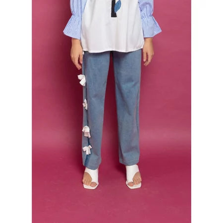 A person standing against a purple background, wearing blue denim pants with decorative white flowers on the leg, paired with a white shirt and beige shoes.

Seorang wanita berdiri dengan latar belakang ungu, mengenakan celana denim biru dengan hiasan bunga-bunga putih di bagian kaki, dipadukan dengan kemeja putih dan sepatu krem.