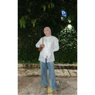 A person wearing a white vest by Abame with a floral pattern, blue jeans, and brown shoes, standing on a sidewalk with greenery in the background.