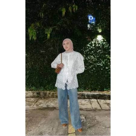 A person wearing a white vest by Abame with a floral pattern, blue jeans, and brown shoes, standing on a sidewalk with greenery in the background.