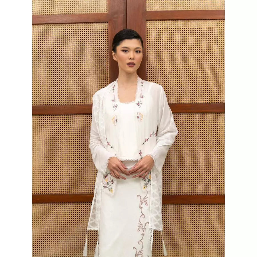 Woman wearing a white embroidered kebaya by Kartinis Label standing against a woven wall.