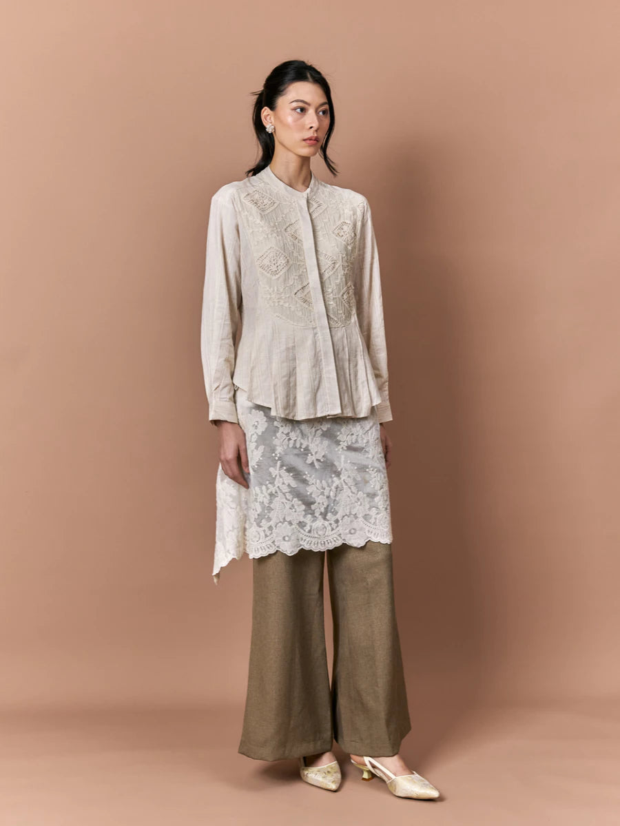 Woman wearing a beige embroidered  shirt by Studio Tui, lace skirt, and brown pants against a beige background