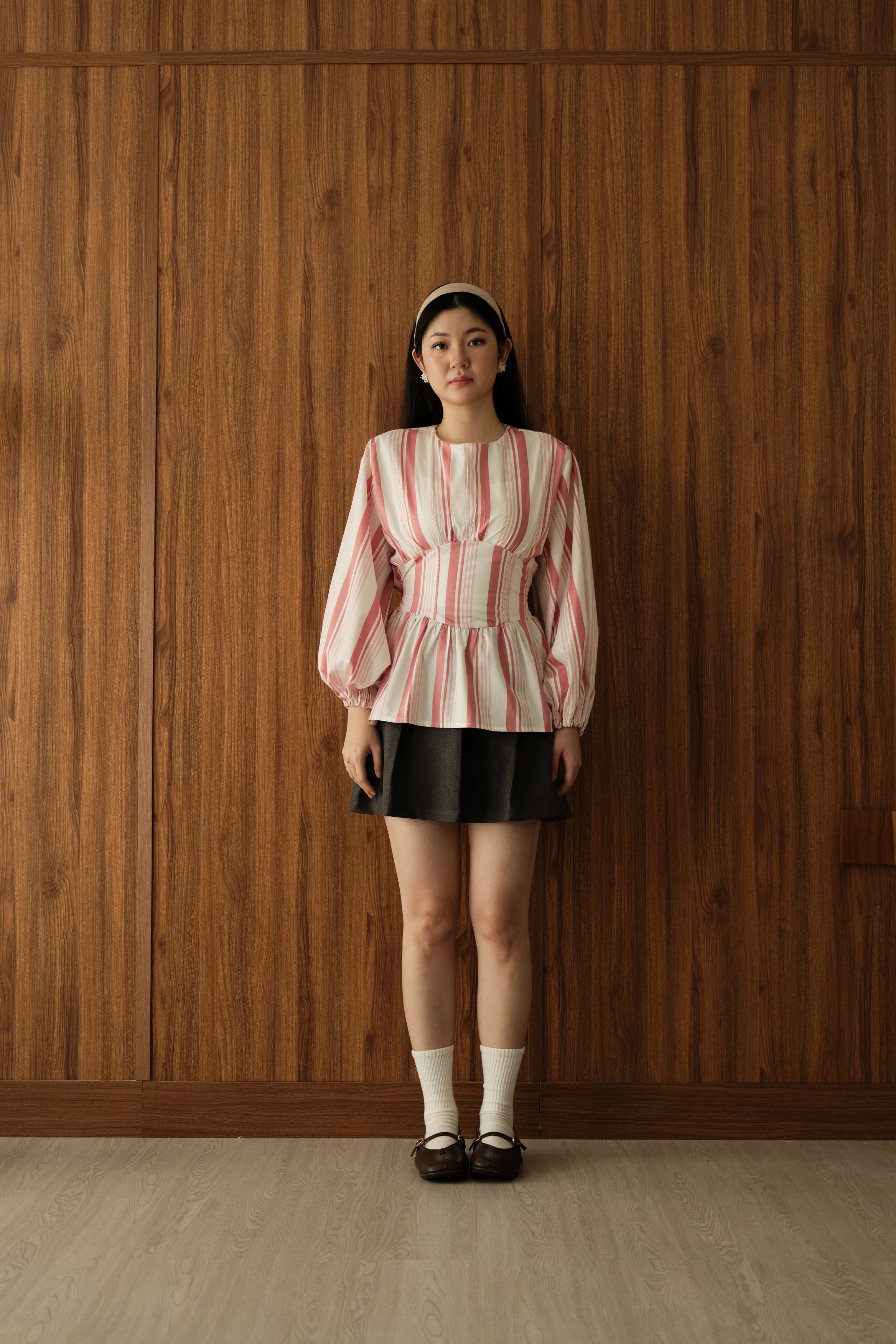 A young woman stands against a wood-paneled wall wearing a pink and white vertically striped long-sleeve blouse by BWBYAZ with a peplum waist, a black pleated mini skirt, white socks, and dark brown Mary Jane shoes.