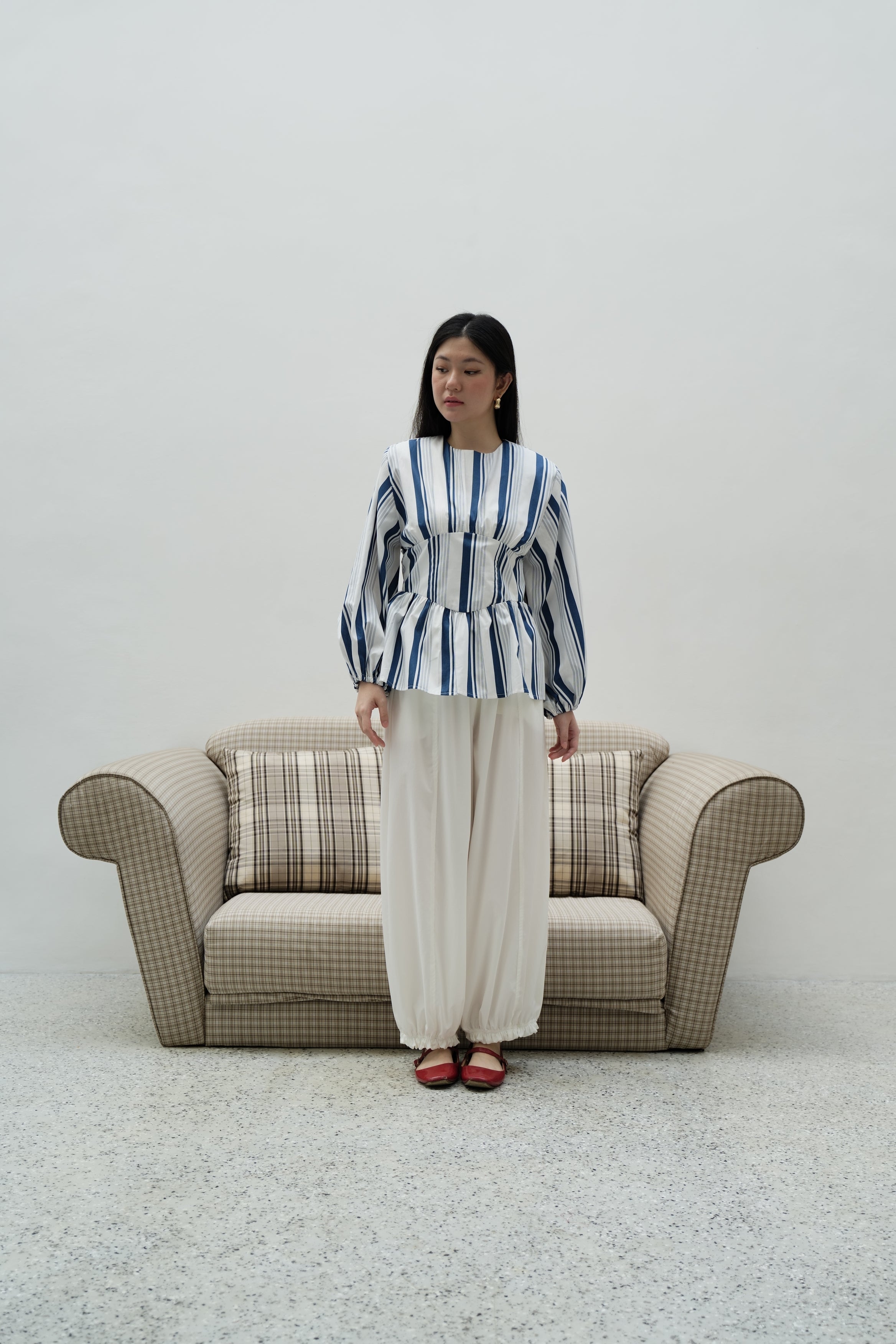 A woman stands in front of a beige armchair wearing a blue and white striped long-sleeve peplum top by BWBYAZ and white wide-leg pants, with red shoes, against a plain light gray wall.