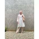 A woman standing outdoors wearing a cream-colored tunic with long sleeves, round neckline, and long pants. She has a bag and is posing against a gray wall with a textured background.