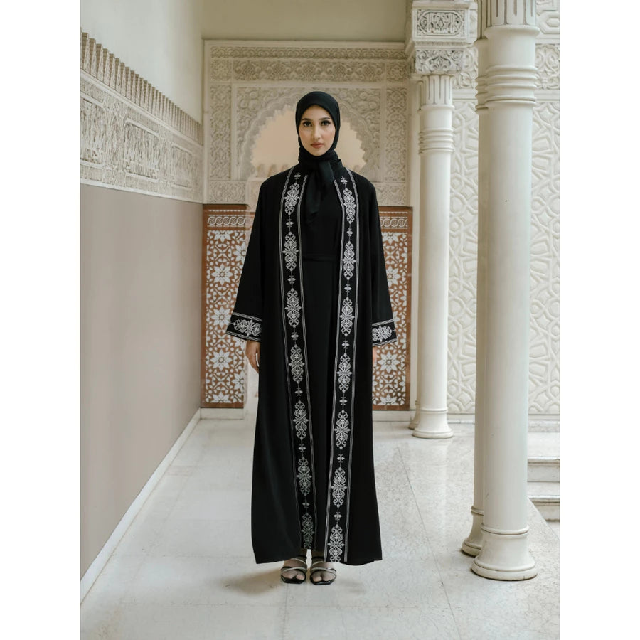 Woman in a black abaya with white embroidery by Tubita standing in an ornate hallway.