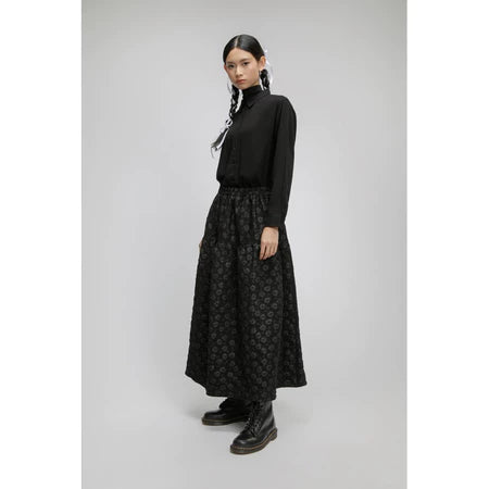 Person wearing a black turtleneck and patterned long black skirt by Yestoday on a gray background