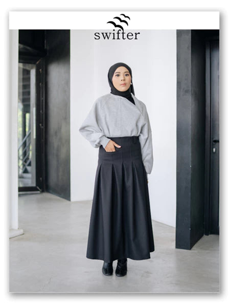 Woman wearing a gray sweater and black pleated skirt by Swifter with a hijab, standing indoors.