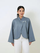 A woman wearing a slate blue cropped blouse by Studio Tui with long wide sleeves and a small white embroidered detail on the chest, paired with white pants, against a plain off-white background.