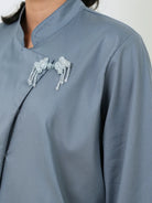 Close-up of a person wearing a light blue-gray long-sleeve blouse by Studio Tui with a mandarin collar and decorative silver beaded brooches on the chest.